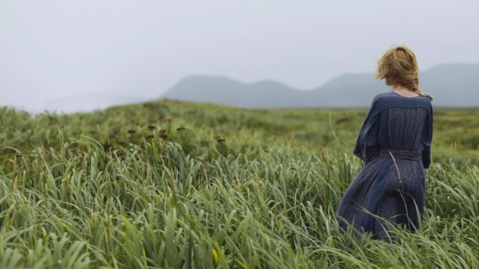 A woman standing in the middle of a field of long green grass with a mountain in the background.